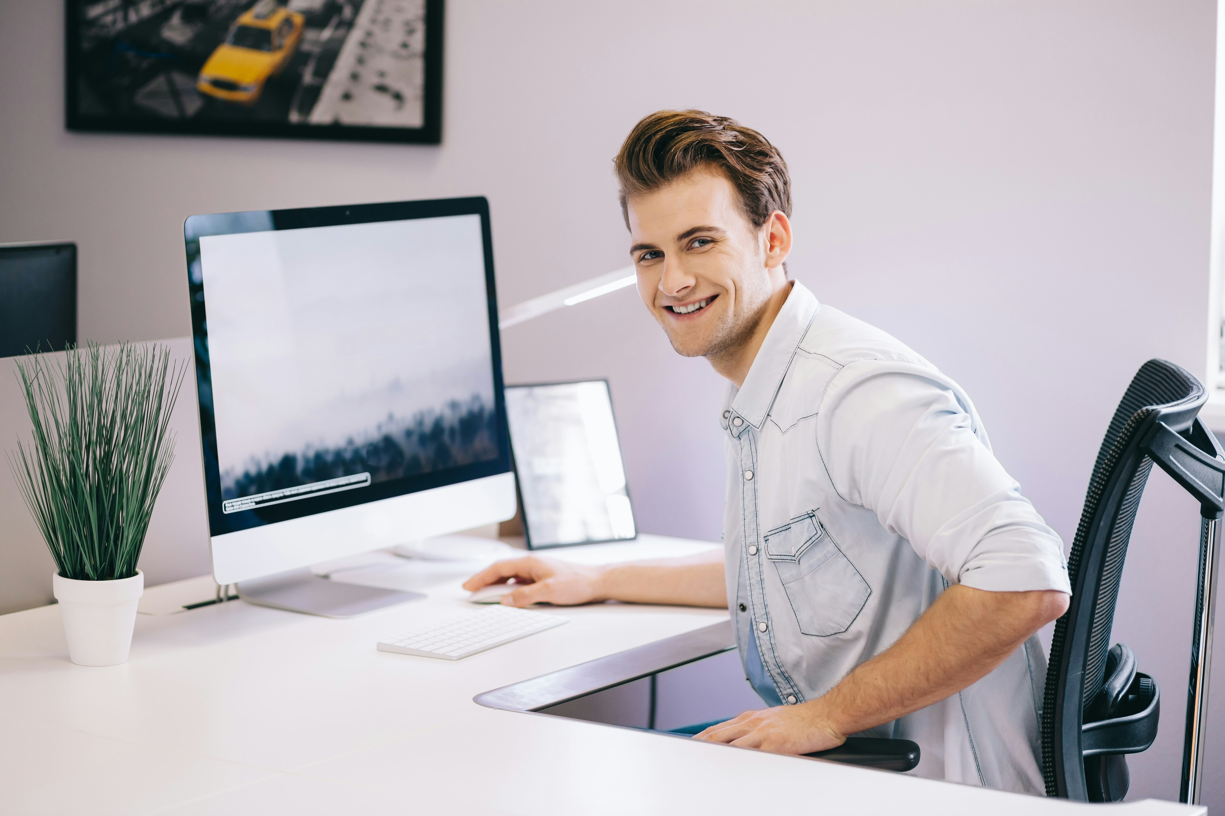 Young worker sitting in an office at the computer. Freelancer in a white shirt. The designer sits in front of a window in the workplace.