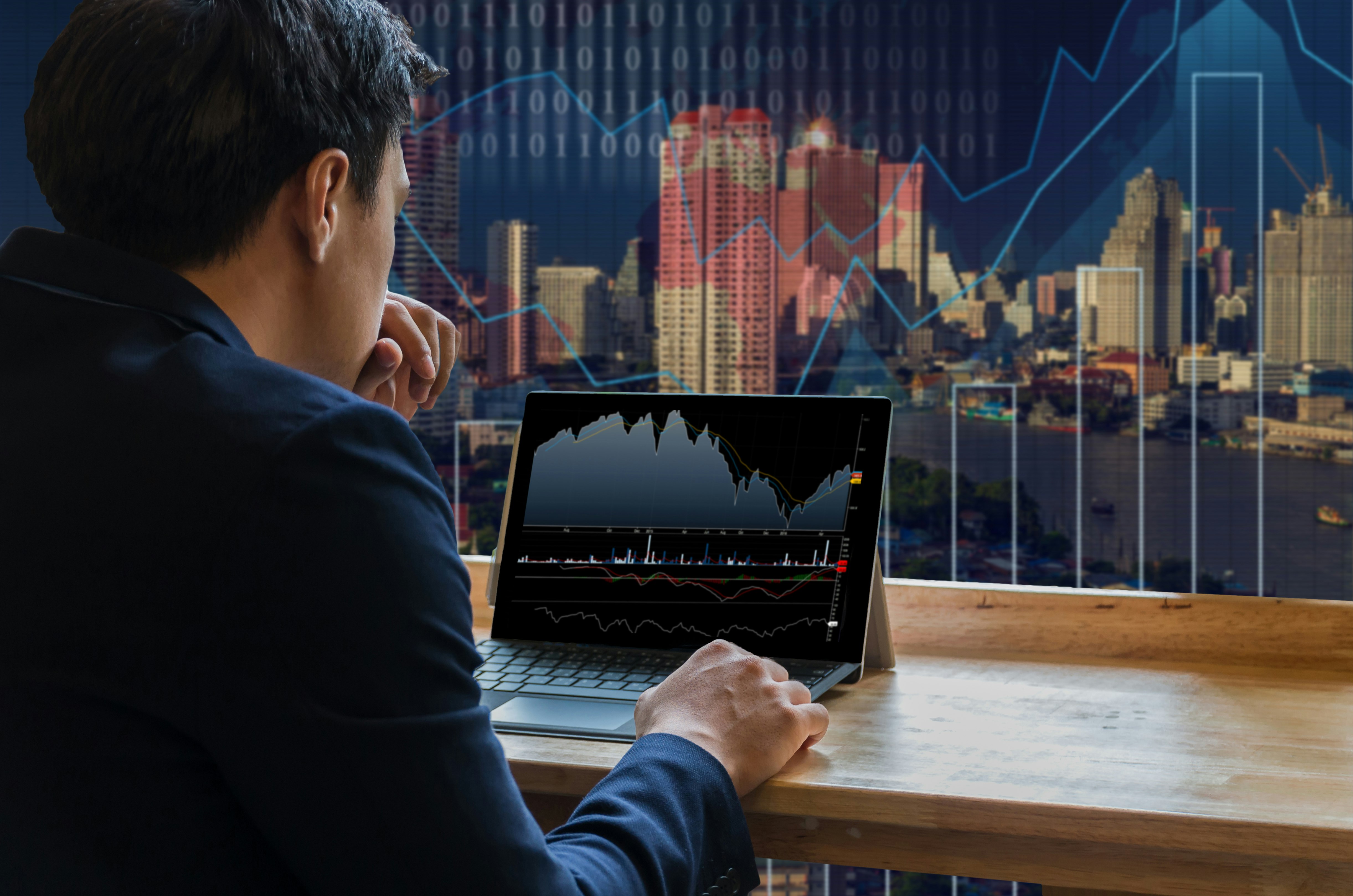 Businessman sitting and using computer laptop showing trading graph beside the windows glass over the Trading graph on the cityscape background, Business financial concept